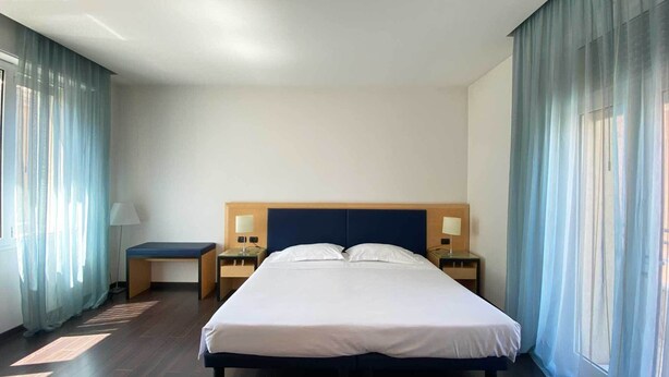 Una camera d'hotel con un letto grande, due comodini con lampade e tende leggere alle finestre.