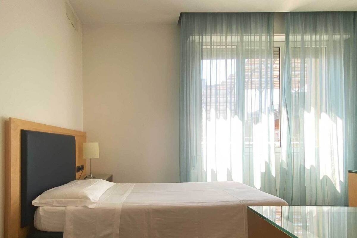 Une chambre d’hôtel avec un lit, une table de chevet avec une lampe, une table en verre et une vue à travers des rideaux transparents.