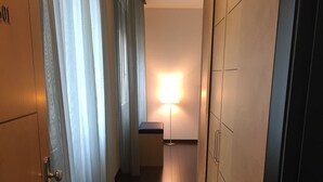 Un corridoio stretto con una porta in legno a sinistra, una lampada da terra al centro e una porta scura a destra.