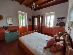 Quarto com cama de madeira, duas janelas, um espelho e um suporte para panelas suspenso.