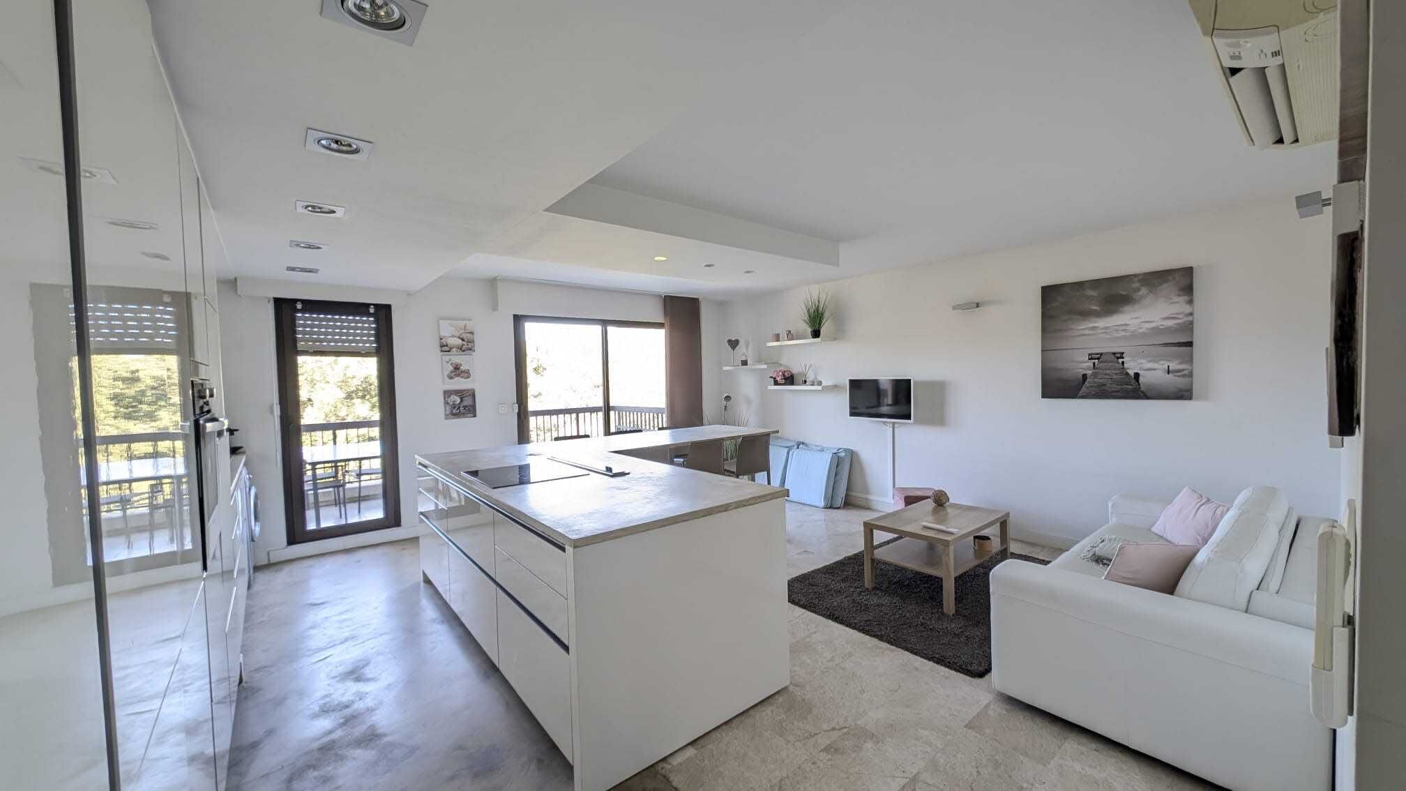 Une cuisine moderne avec un îlot central, un coin repas avec une table et des chaises, un salon avec un canapé et un balcon offrant une vue.
