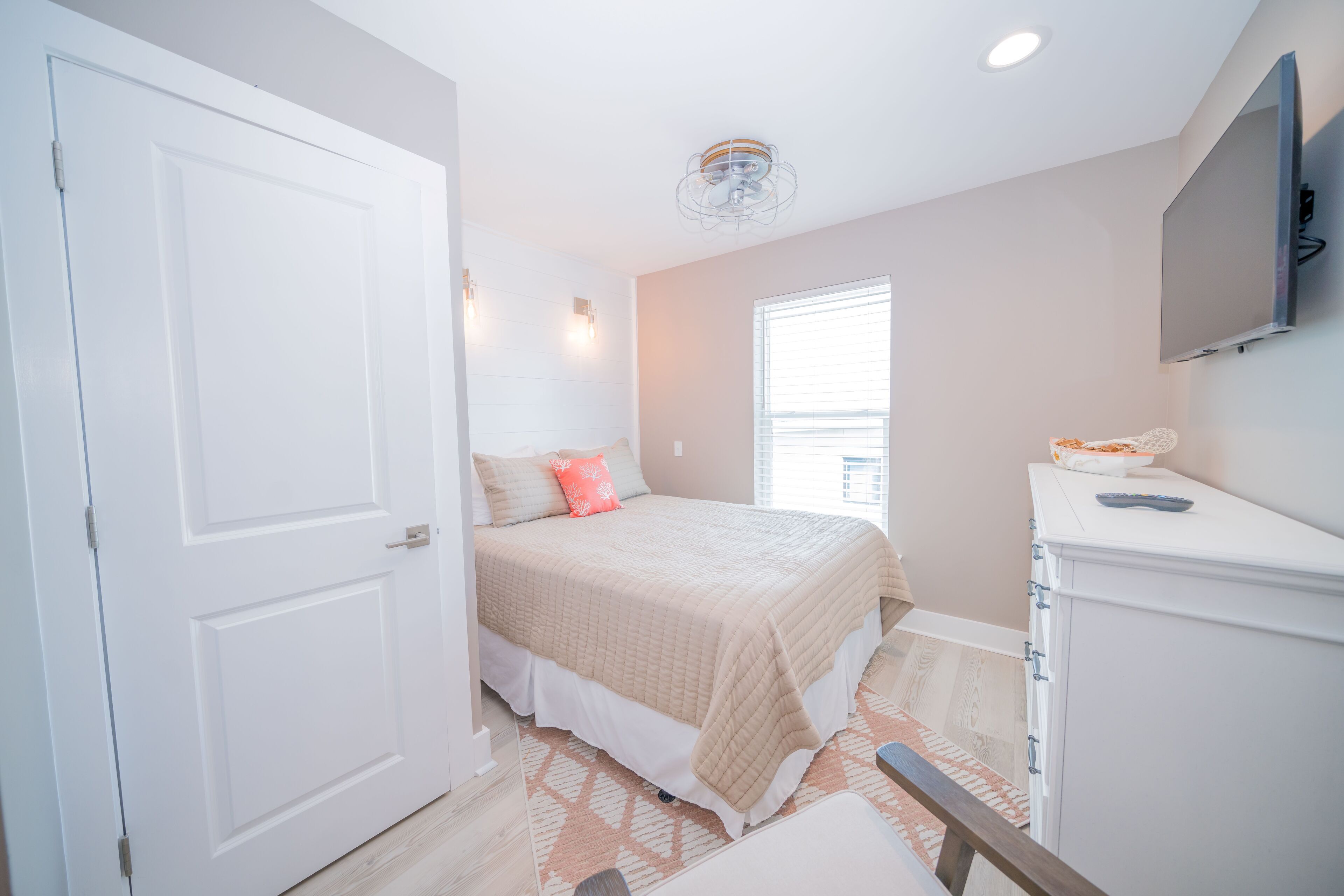 A bedroom with a bed, a dresser, a TV mounted on the wall, and a window with blinds.