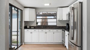 A modern kitchen with white cabinets, a stainless steel refrigerator, and a window overlooking a tree. - Clearwater Game House! Heated Pool, Bowling, Golf! (Largo)