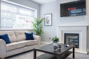 Un salon avec un canapé beige, une table basse, une cheminée, un téléviseur fixé au mur et une grande fenêtre avec des stores.