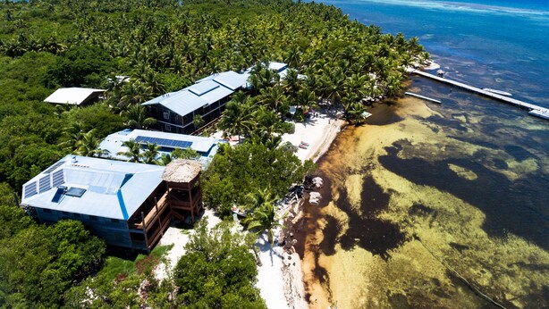 An aerial view of a tropical resort with multiple buildings, a beach, and a pier extending into the ocean. - Itza Resort (Lighthouse Reef)