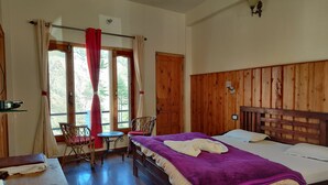 A bedroom with a wooden headboard, a bed with a purple blanket, a small table with chairs, and a window with curtains. - Hotel Burans And Restaurant (Chakrata)