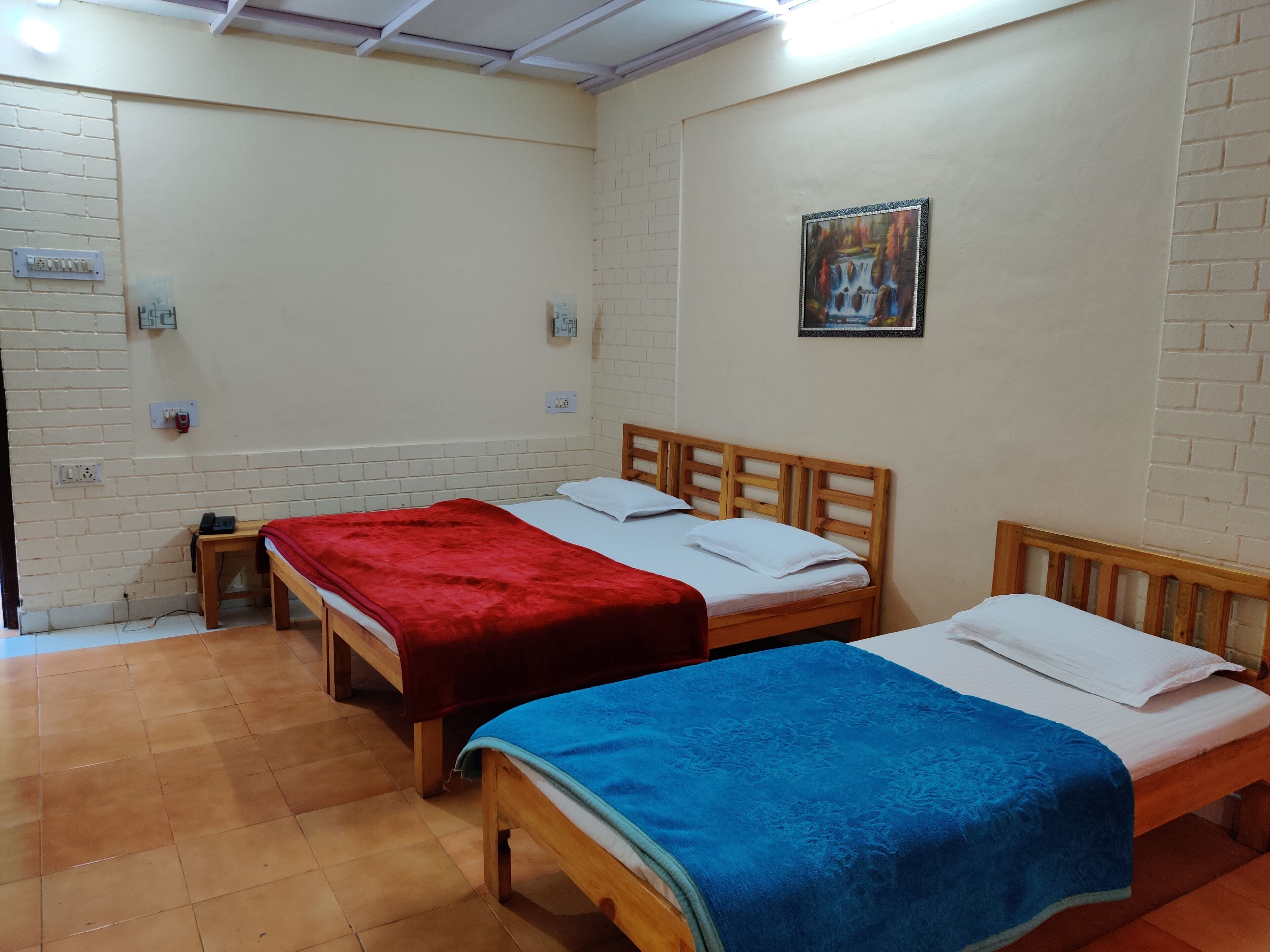 Una habitación con dos camas de madera, cada una con colchón blanco y manta roja y azul. Hay una mesita con un objeto rojo encima, y un cuadro colgado en la pared.