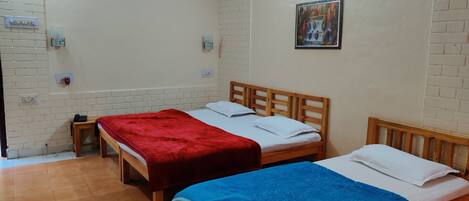 Una habitación con dos camas de madera, cada una con colchón blanco y manta roja y azul. Hay una mesita con un objeto rojo encima, y un cuadro colgado en la pared.