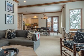 Un vaste espace de vie avec un canapé gris, des coussins à motifs et une table basse. On y trouve également un coin repas avec une table et des chaises en bois, et une cuisine à l’arrière-plan.