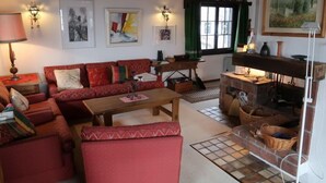 A cozy living room with a red sofa set, a wooden coffee table, and a fireplace. There are framed artworks on the walls and a window with green curtains.