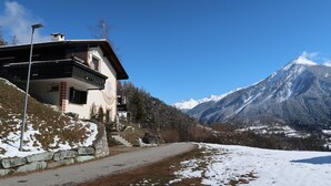 A mountain house with a balcony, a stone wall, and a snow-covered path leading to a mountain.