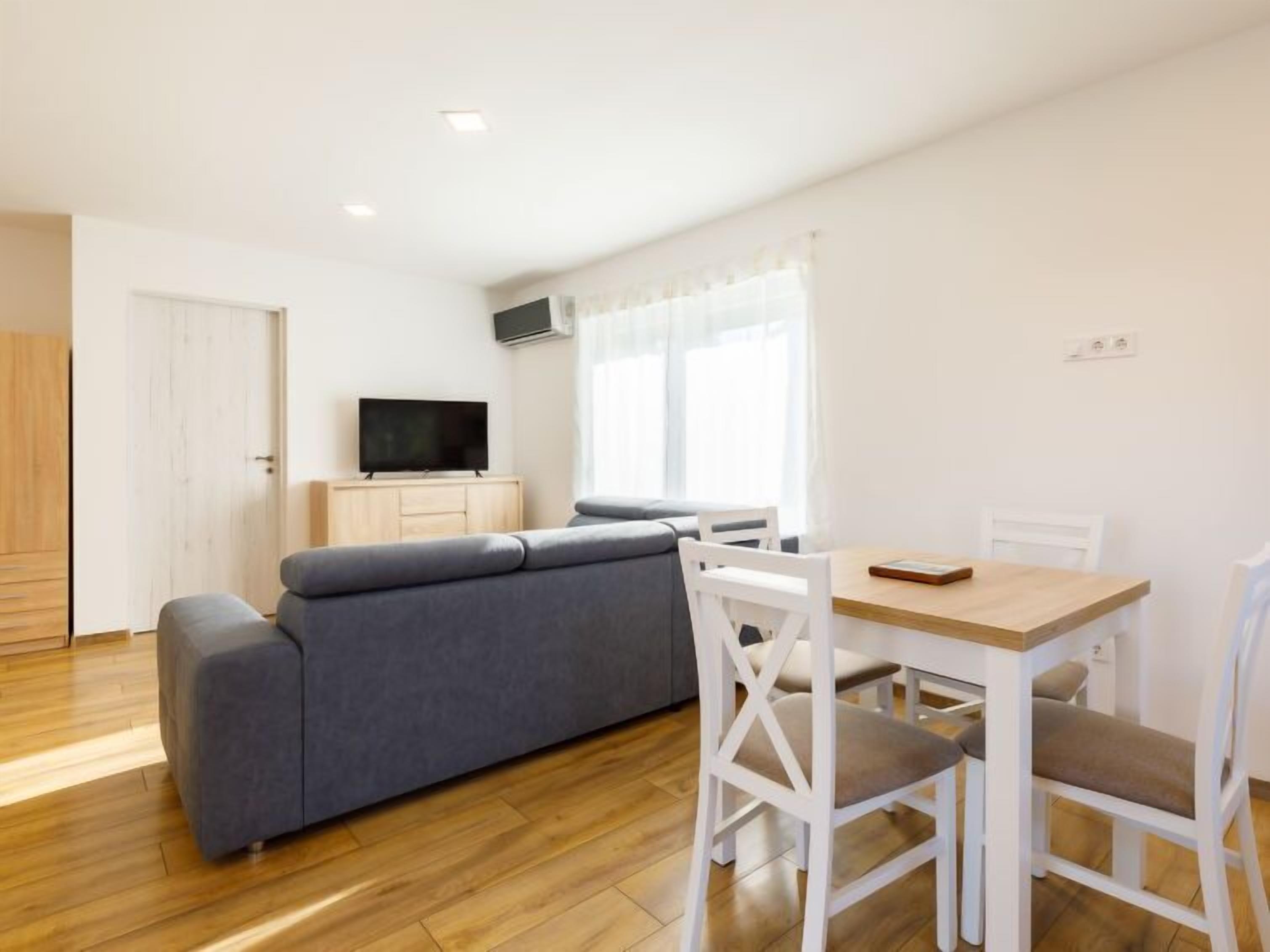 Un salon moderne avec un canapé gris, des chaises blanches, une table à manger en bois et un téléviseur à écran plat posé sur un meuble en bois.