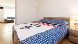 Ein Schlafzimmer mit Bett, Holz-Kopfteil, Nachttisch, Lampe und Blick auf einen Wohnbereich mit Sofa und Fenster.