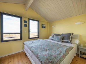 A bedroom with a large bed, a nightstand, a window with a view of the beach, and a ceiling with wooden beams.