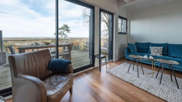 Een ruime woonkamer met een grote schuifpui naar een terras, een leren bank, een blauwe zithoek en een kleine ronde tafel.