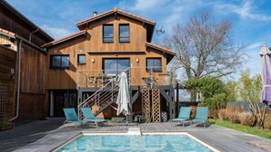 Een houten, tweelaags huis met een zwembad, buitenmeubels en een trap naar een balkon.