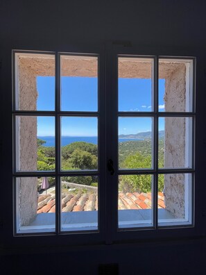 Una stanza con una grande finestra divisa in nove vetri, che si affaccia su un paesaggio costiero con vegetazione e una cittadina in lontananza.