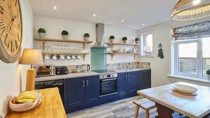 A modern kitchen with blue cabinets, a wooden table, and a window with blinds. - Host & Stay | Mumbles Cottage (Whitby)