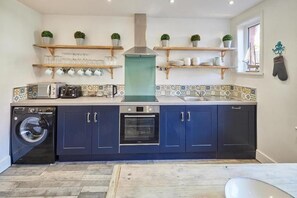 A compact kitchen with dark blue cabinets, a built-in oven, a washing machine, and a tiled backsplash. - Host & Stay | Mumbles Cottage (Whitby)
