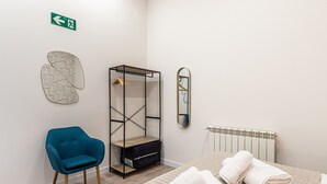 Ein Zimmer mit blauem Stuhl, einem Metallregal, einem Spiegel und einem Bett mit gerollten Handtüchern.