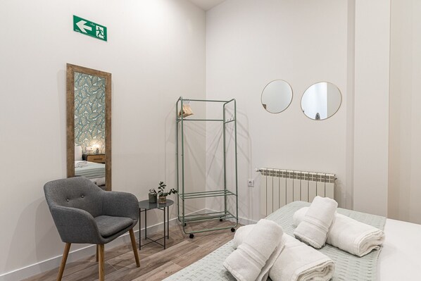 A hotel room with a bed, a chair, a radiator, a mirror, and a green exit sign.