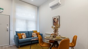 Ett modernt vardagsrum med en blå soffa, orangefärgade stolar, ett matbord för fyra personer och ett stort fönster med tunna, genomskinliga gardiner.