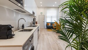 Ett modernt kök med en inbyggd mikrovågsugn, en diskho och en kaffemaskin. I vardagsrummet finns en soffa och ett soffbord, och i hörnet står en stor grön växt.