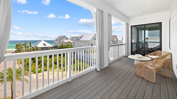 A balcony with a view of the beach and houses, wicker furniture, and a glass door.