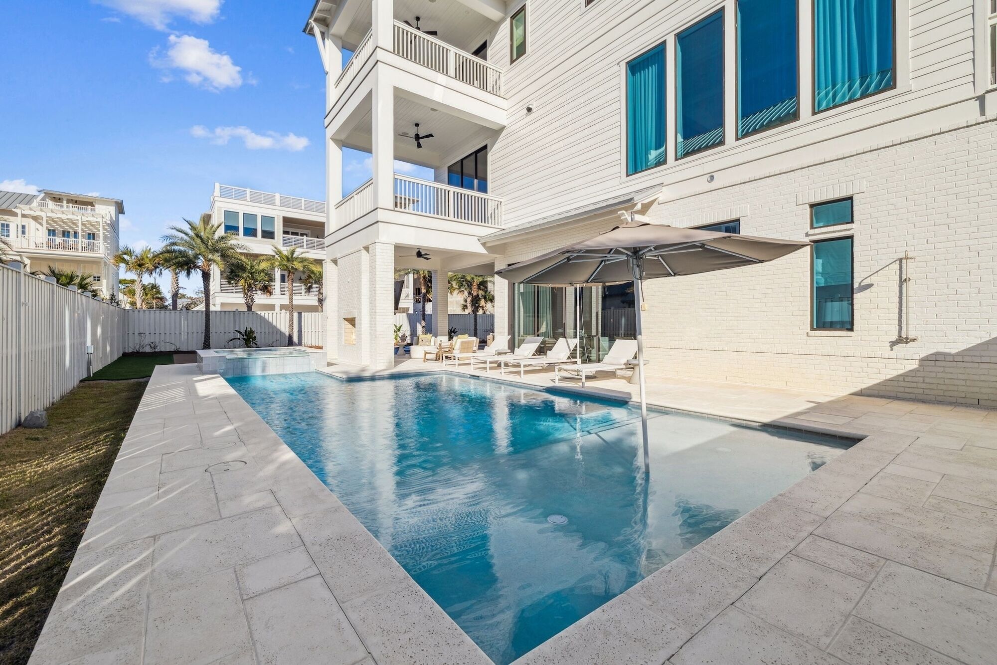 A swimming pool with lounge chairs and umbrellas, surrounded by a white building with balconies and large windows.