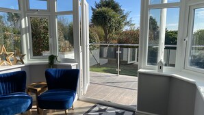 A room with a bay window overlooking a garden, featuring two blue chairs, a small table, and a star-patterned doormat. - Willows Worthing 4 bed With ev Charger & sea Views (Worthing)