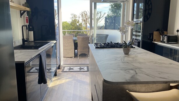 A modern kitchen with a marble countertop, a gas stove, and a dining area with wicker chairs. - Willows Worthing 4 bed With ev Charger & sea Views (Worthing)