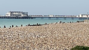 A coastal scene with a pebbly beach, a pier extending into the water, and a building with multiple floors. - Willows Worthing 4 bed With ev Charger & sea Views (Worthing)