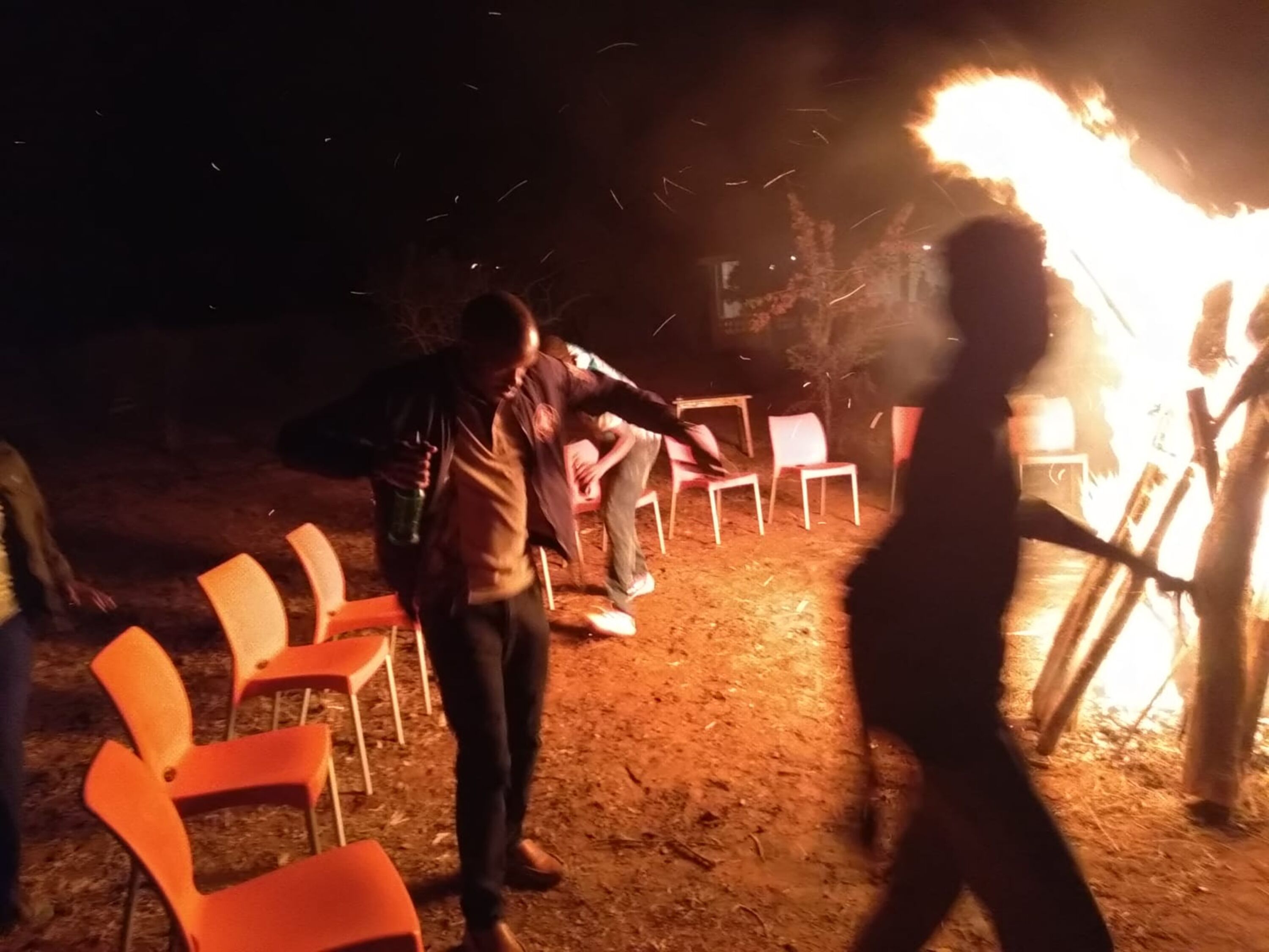 Um grupo de pessoas reunido em volta de uma fogueira em ambiente externo, com cadeiras dispostas em semicírculo.