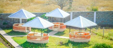 A series of outdoor seating areas with round tables and chairs, surrounded by decorative orange lattice walls, set against a backdrop of a stone wall and grassy field.