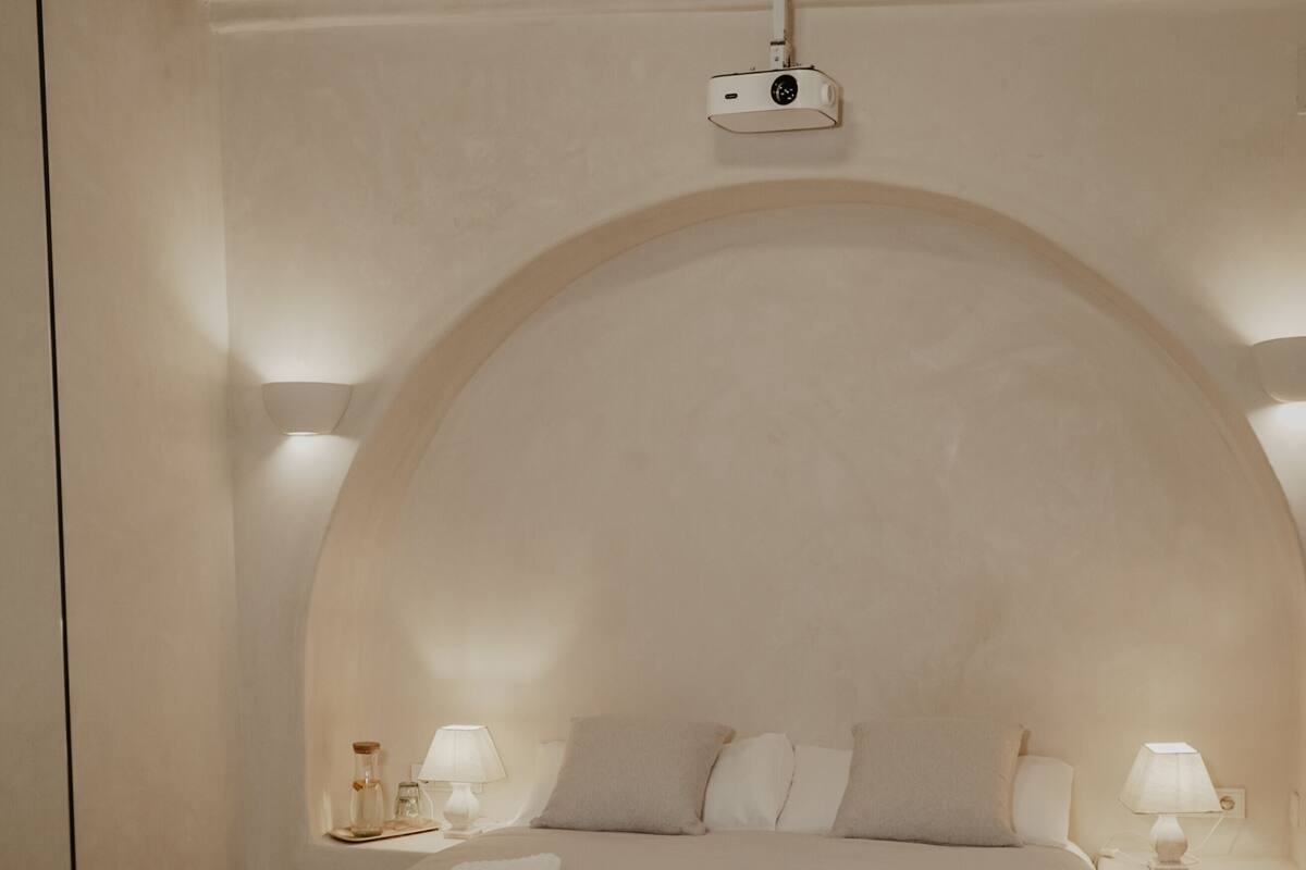 Un dormitorio con cama, lámparas de noche, una mesita y un proyector montado en la pared.