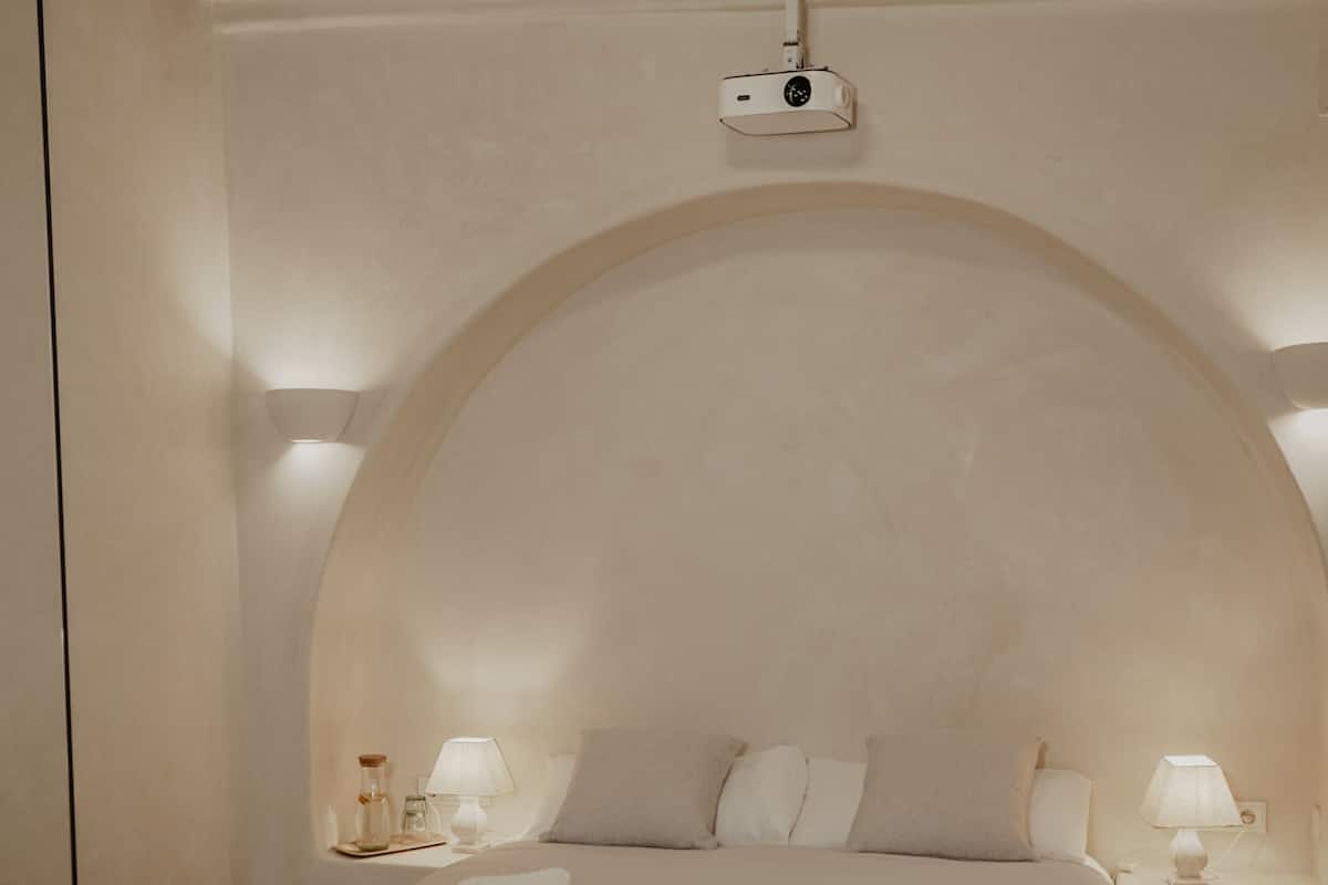 A bedroom with a bed, bedside lamps, a small table, and a projector mounted on the wall.