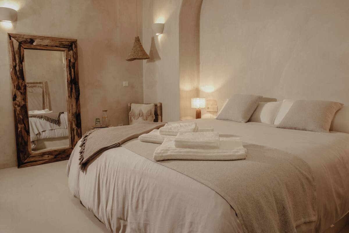 Un dormitorio con una cama grande, un espejo con marco de madera, una mesita de noche con lámpara y un aplique de pared.