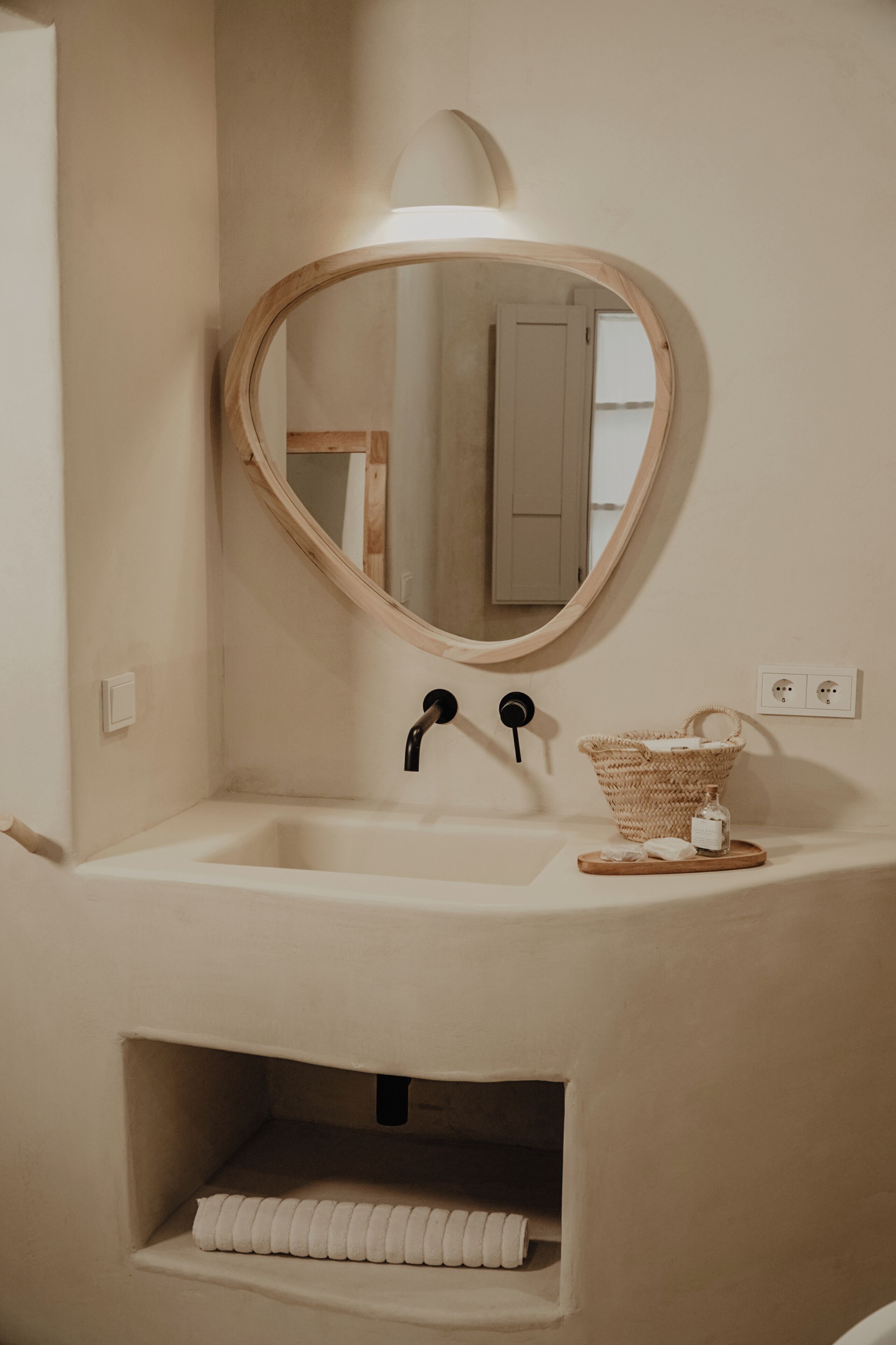 Un baño con un espejo redondo, un lavamanos, una cesta y un perchero para toallas.