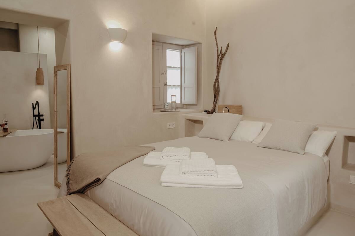 Un dormitorio moderno con una cama grande, una bañera integrada, un tocador con espejo y una ventana con cortinas.