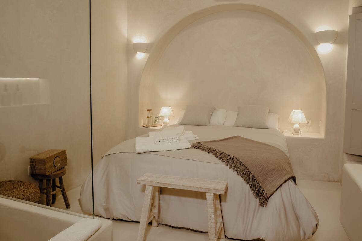 Un dormitorio con una cama grande, dos lámparas de noche, un taburete de madera y un banco.