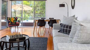 A modern living room with a chessboard, a sofa, a dining table with chairs, and a large window with a view of greenery. - Moonah Link Views, Spa, Sunset, Linen, Wi-Fi, private and serene (Fingal)