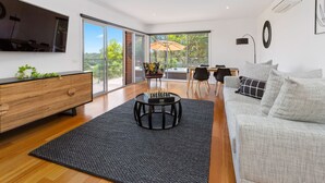 A modern living room with a large TV, a sofa, a coffee table, and a dining area with chairs and a table. - Moonah Link Views, Spa, Sunset, Linen, Wi-Fi, private and serene (Fingal)