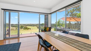 A modern dining area with a long table, black chairs, and a view of a golf course through large windows. - Moonah Link Views, Spa, Sunset, Linen, Wi-Fi, private and serene (Fingal)
