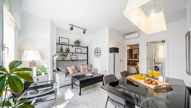 A modern living room with a sofa, coffee table, and dining area. There is a bookshelf with decorative items and a clock on the wall.