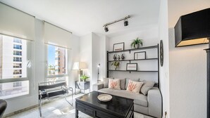 A living room with a black coffee table, a grey sofa, and a black metal bench. There is a wall-mounted shelf with decorative items and a lamp.