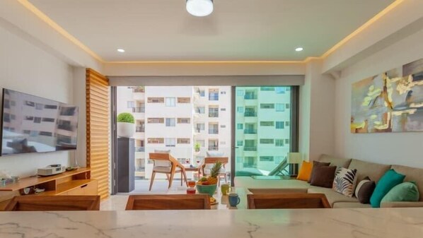 A modern living room with a flat-screen TV, a sofa, a coffee table, and a dining area with chairs and a table. - Incredible new condo 1/2 Block from Beach/Malecon! (Puerto Vallarta)
