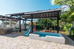 A covered outdoor area with a pool, play structures, and stone flooring.