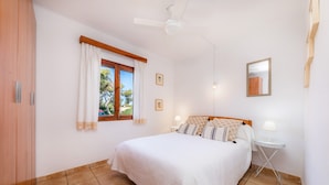 A bedroom with a bed, a ceiling fan, a window with curtains, a bedside table with a lamp, and a wall-mounted mirror.