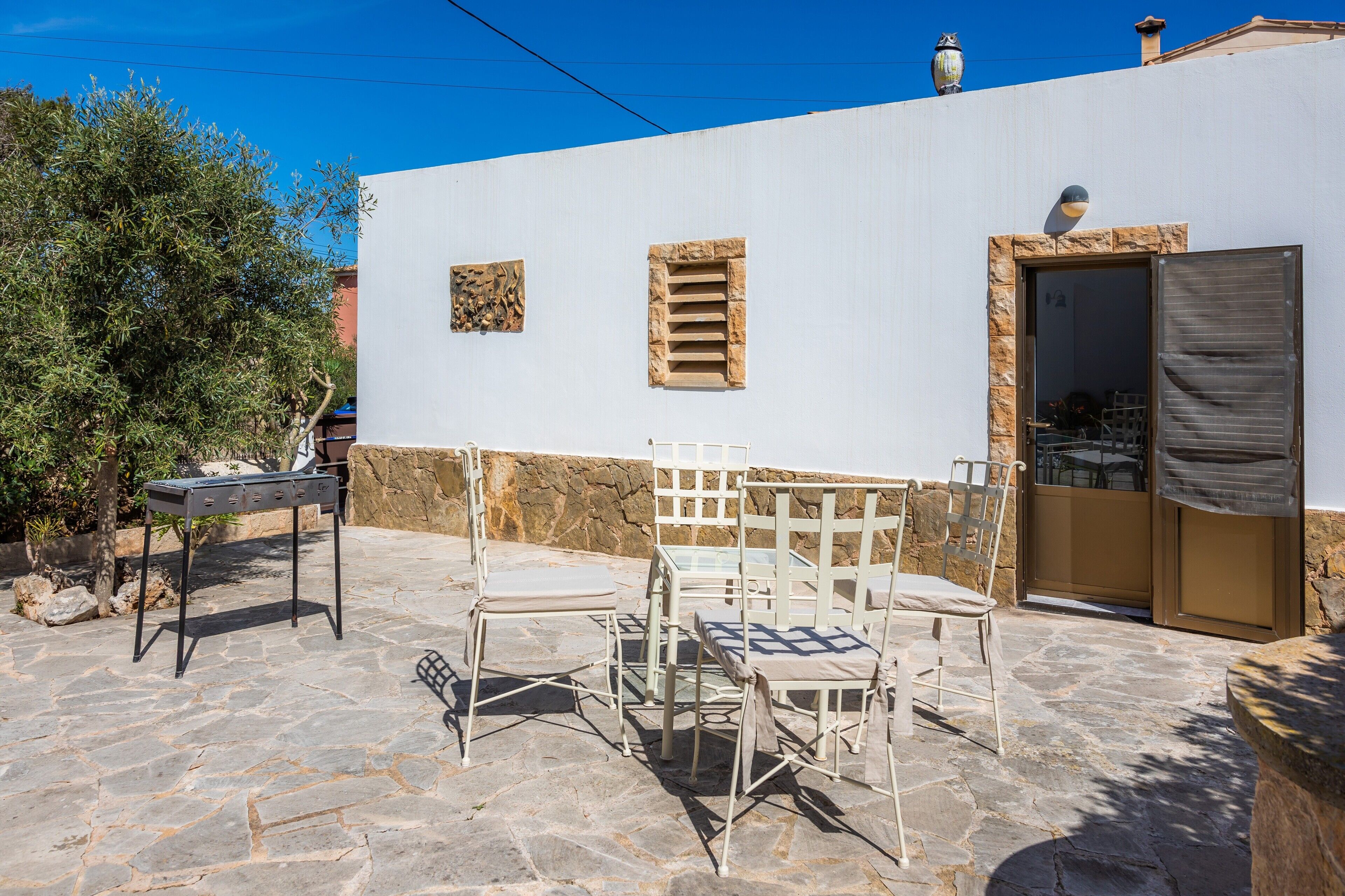 Un patio al aire libre con suelo de piedra, una pared blanca con una puerta, y una mesa con sillas.