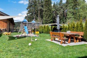 A wooden play structure, picnic table, and benches in a grassy outdoor area with trees in the background.